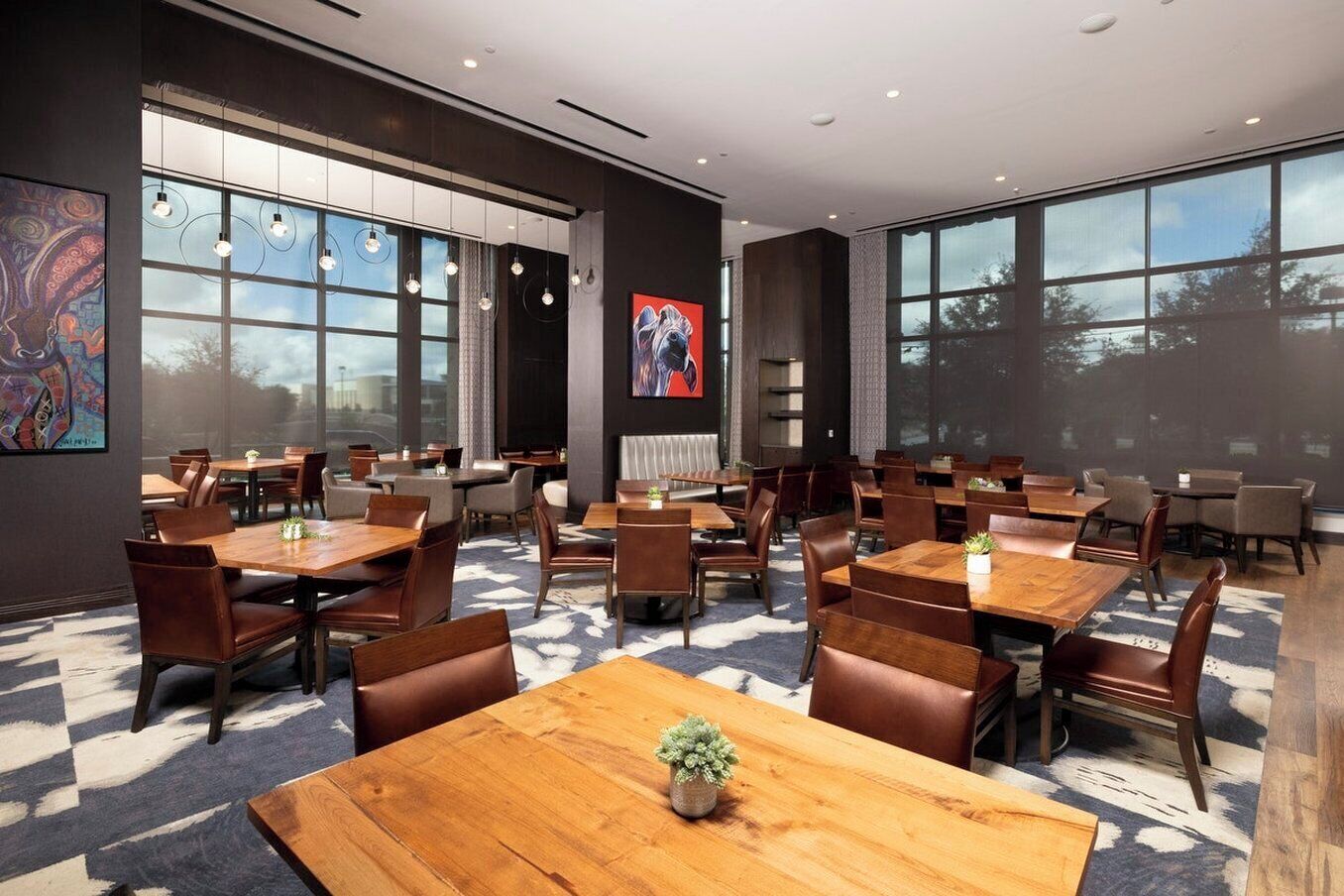 A modern dining area with multiple tables and chairs, large windows, and artwork on the walls.