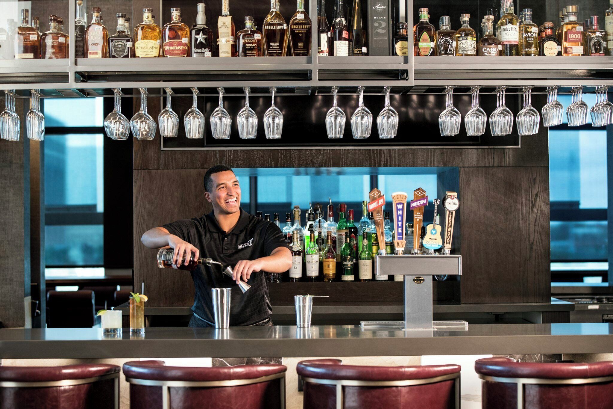 A bartender pouring a drink at a bar with a variety of liquor bottles and a selection of beer taps.