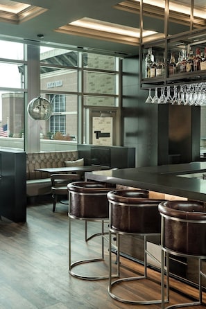 A bar area with leather barstools, a counter, and a glass display case with bottles. - Texas A&M Hotel and Conference Center (College Station)