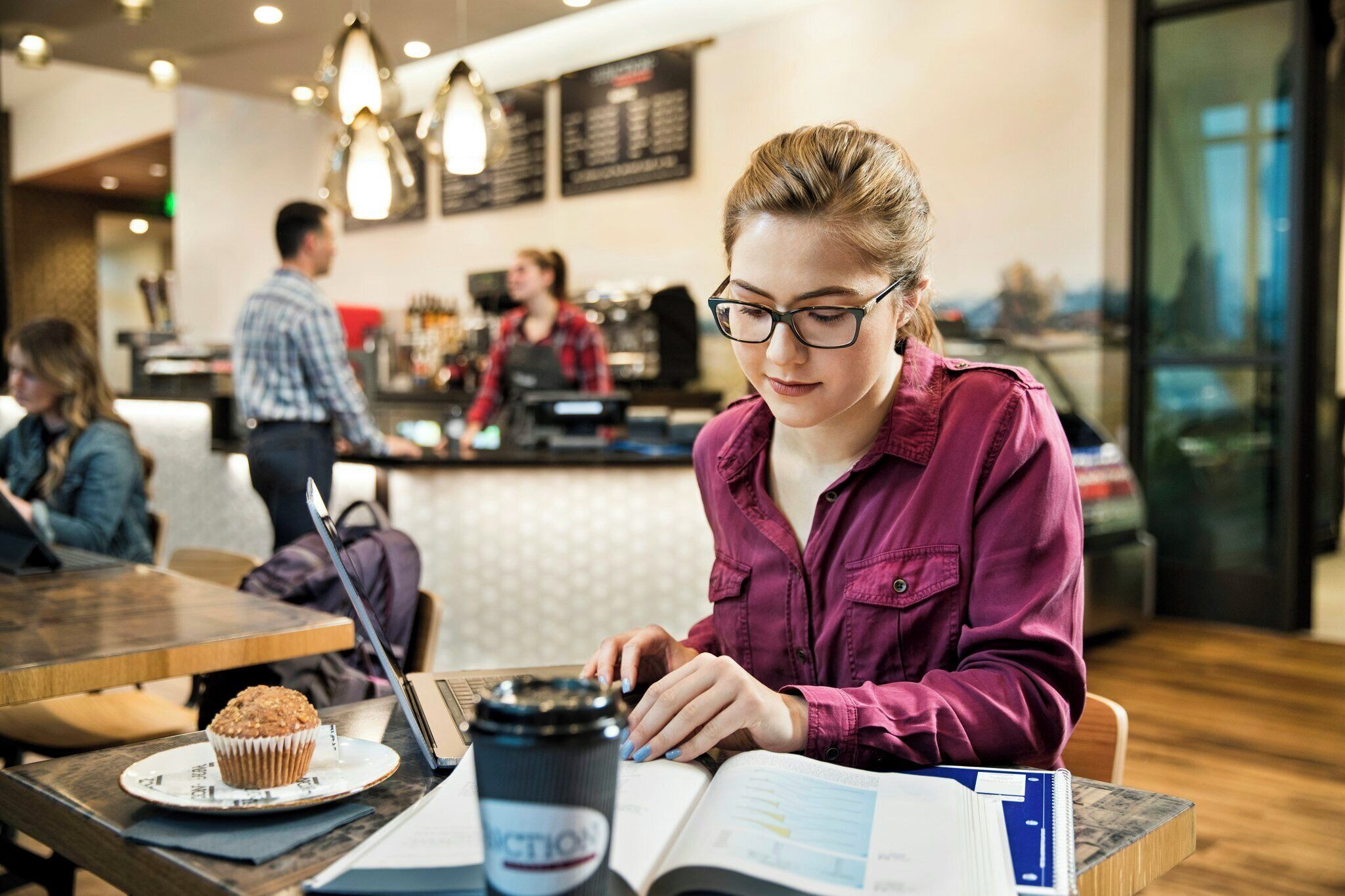 A person sitting at a table with a laptop, a muffin, and a coffee cup, in a cafe with a bar and other patrons in the background.