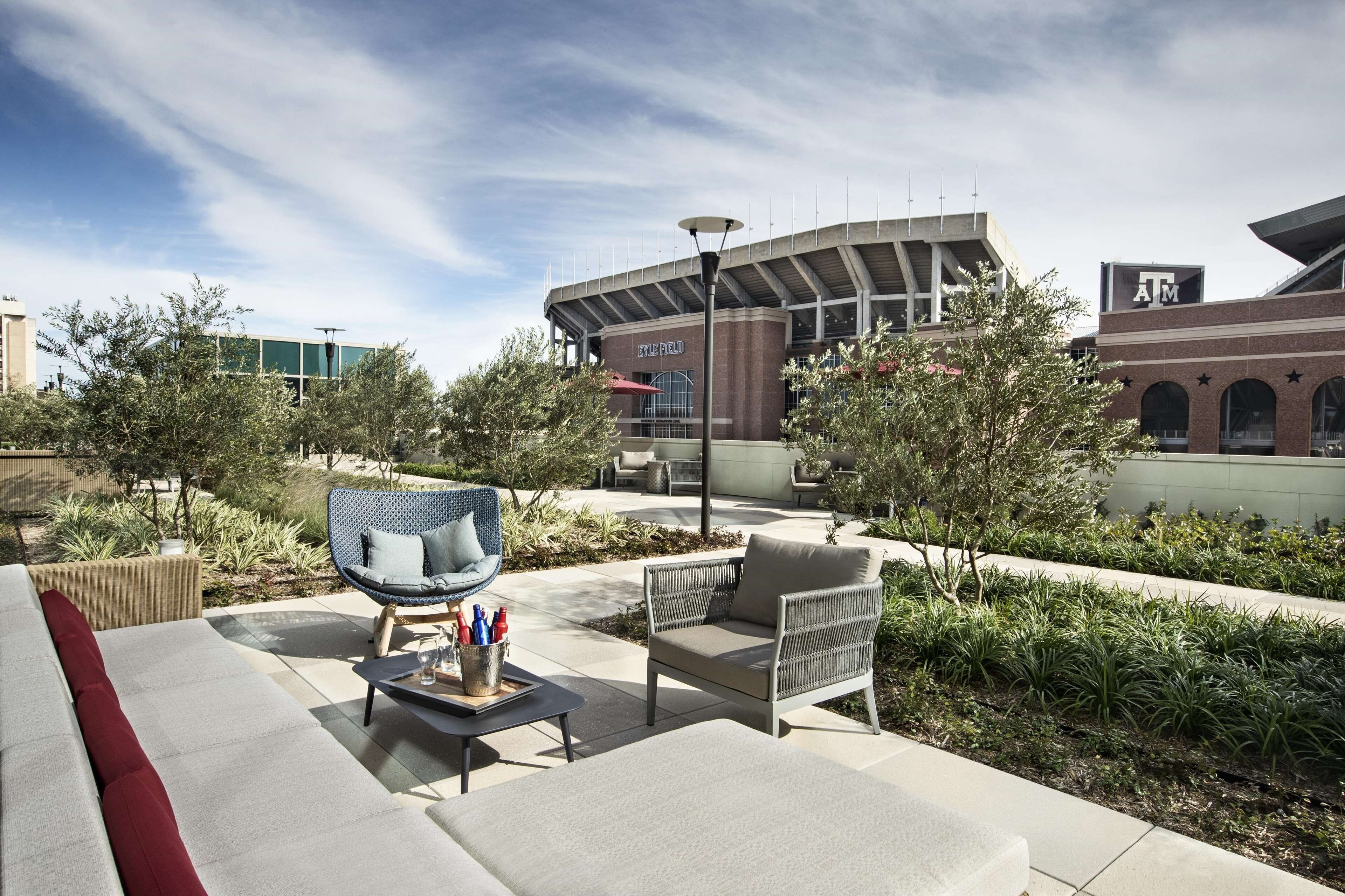 Un área de estar al aire libre con mobiliario moderno, un edificio con la señalización 'Kyle Field' y un cielo despejado.
