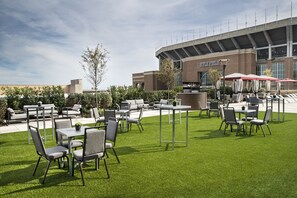 Uma área de estar ao ar livre com mesas e cadeiras, um gramado e um prédio grande com a placa 'Kyle Field' ao fundo.
