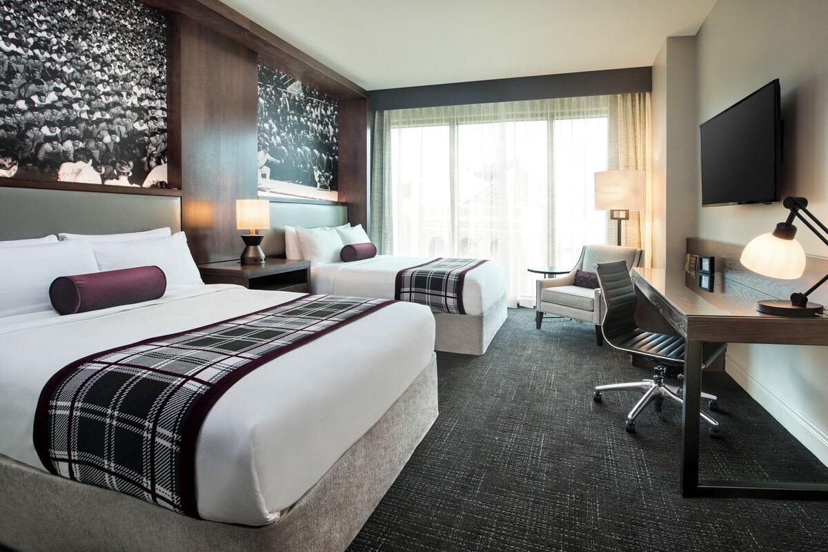 A hotel room with two beds, a desk, a chair, a TV, and artwork on the walls.