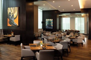 A modern dining area with multiple tables and chairs, large windows, and artwork on the walls. - Texas A&M Hotel and Conference Center (College Station)
