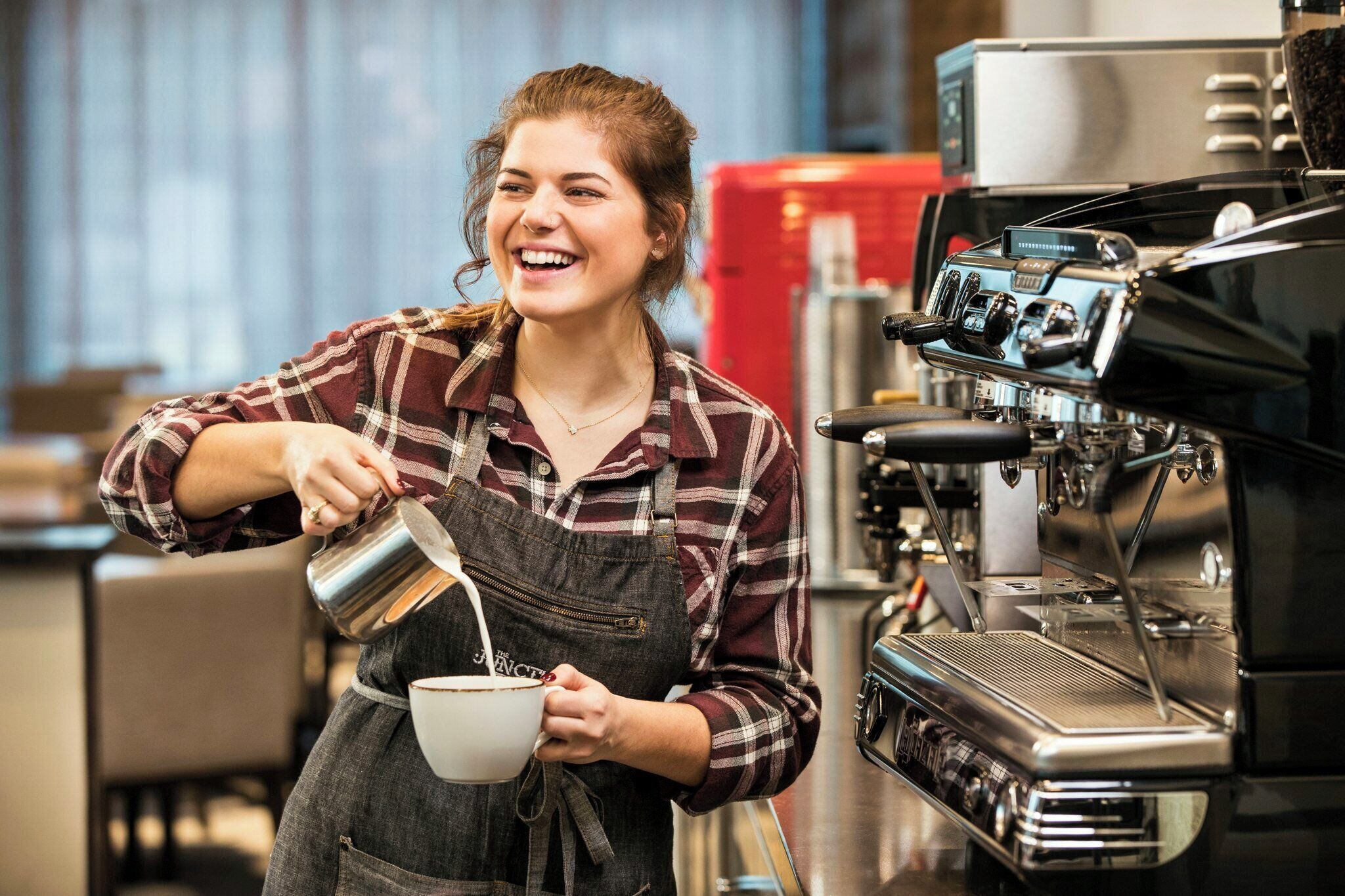 A person pouring milk into a cup, standing in front of a coffee machine.
