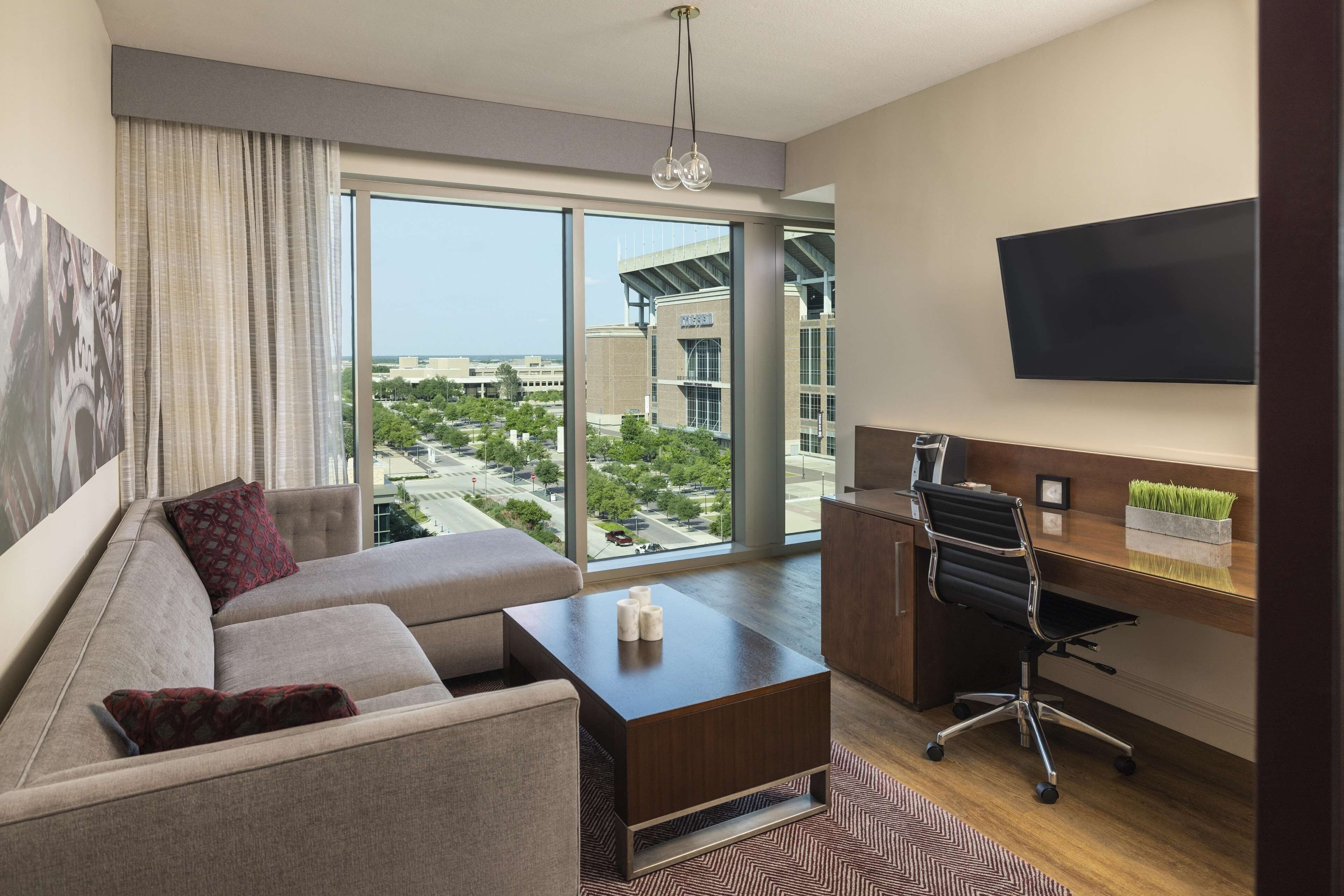 A modern hotel room with a large window, a sofa, a desk, and a flat-screen TV.