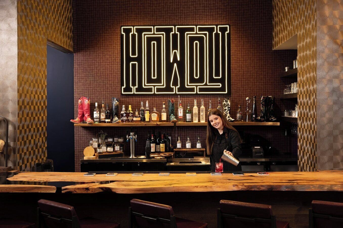 Een bar met een groot verlicht bord waarop 'HOW YOU' staat en planken vol flessen.