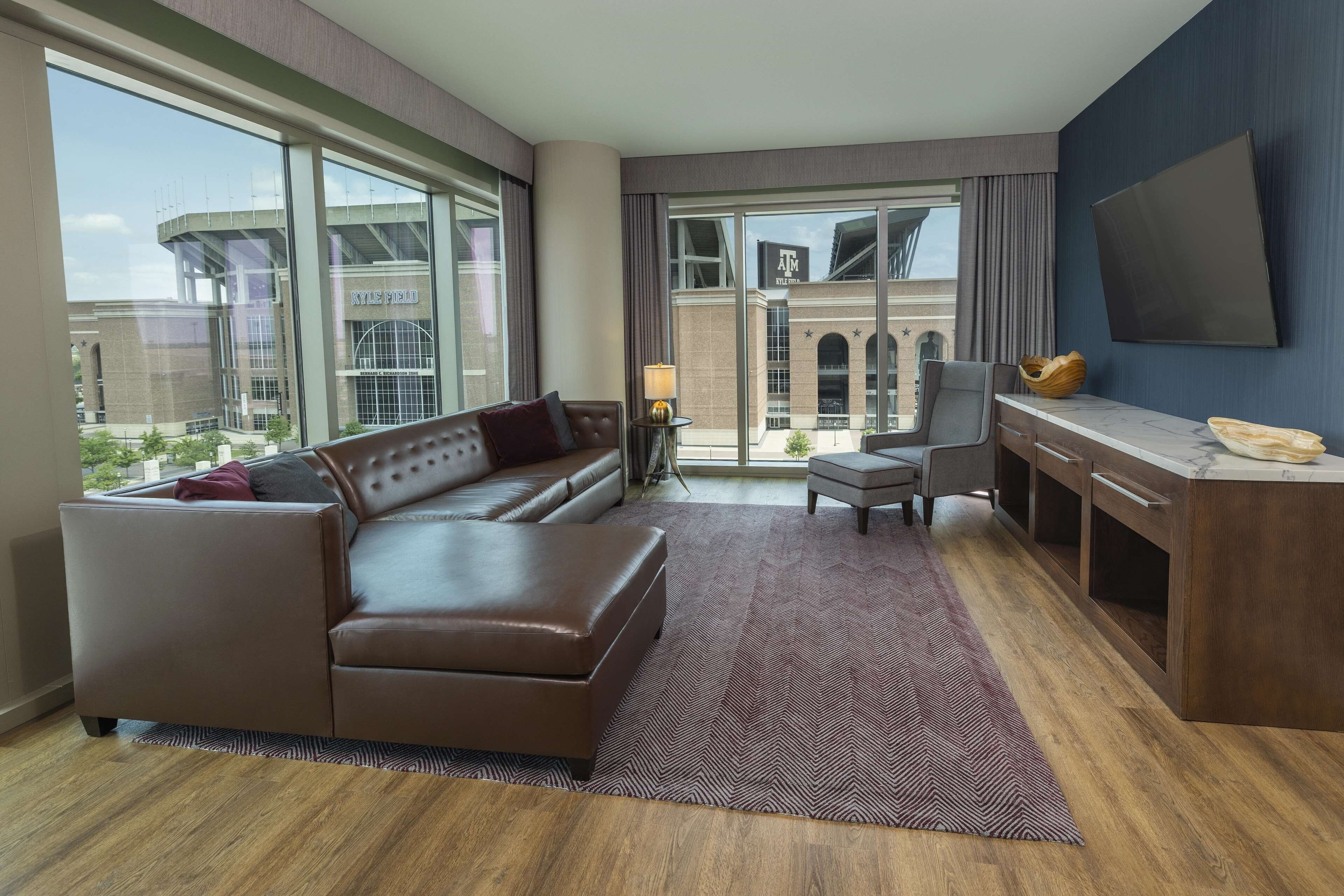 A modern hotel room with a leather sofa, a desk, a chair, and large windows offering a view of the cityscape.
