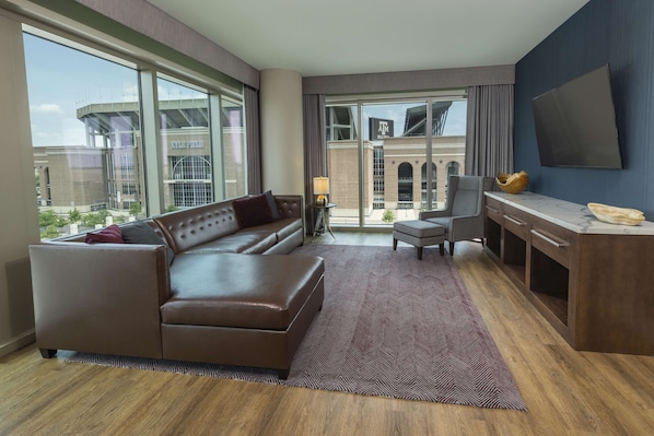 A modern hotel room with a leather sofa, a desk, a chair, and large windows offering a view of the cityscape. - Texas A&M Hotel and Conference Center (College Station)