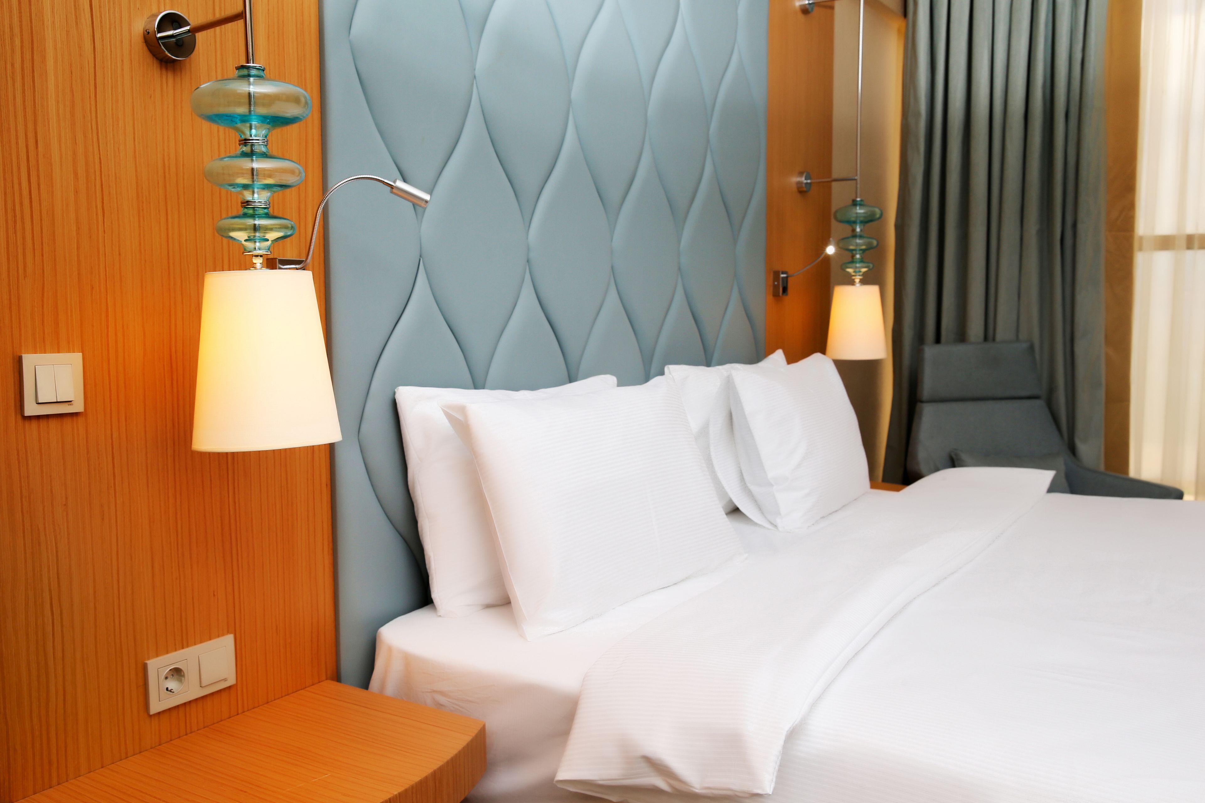 Une chambre d’hôtel avec une tête de lit capitonnée, une table de chevet en bois, un lit avec du linge de lit blanc et deux lampes fixées au mur.