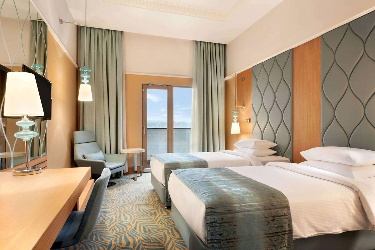 A hotel room with two beds, a desk, a chair, a small table, and a view of the outdoors.