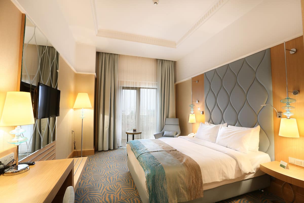 A hotel room with a large bed, two bedside tables, a desk, a chair, a TV, and a window with curtains.