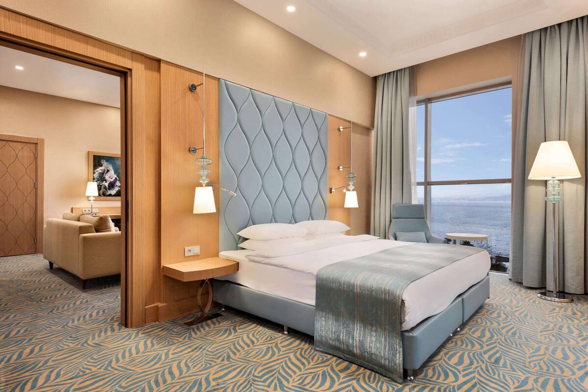 A hotel room with a large bed, a wooden headboard, a nightstand, a desk, a chair, a view of the sea, and a large window with curtains.