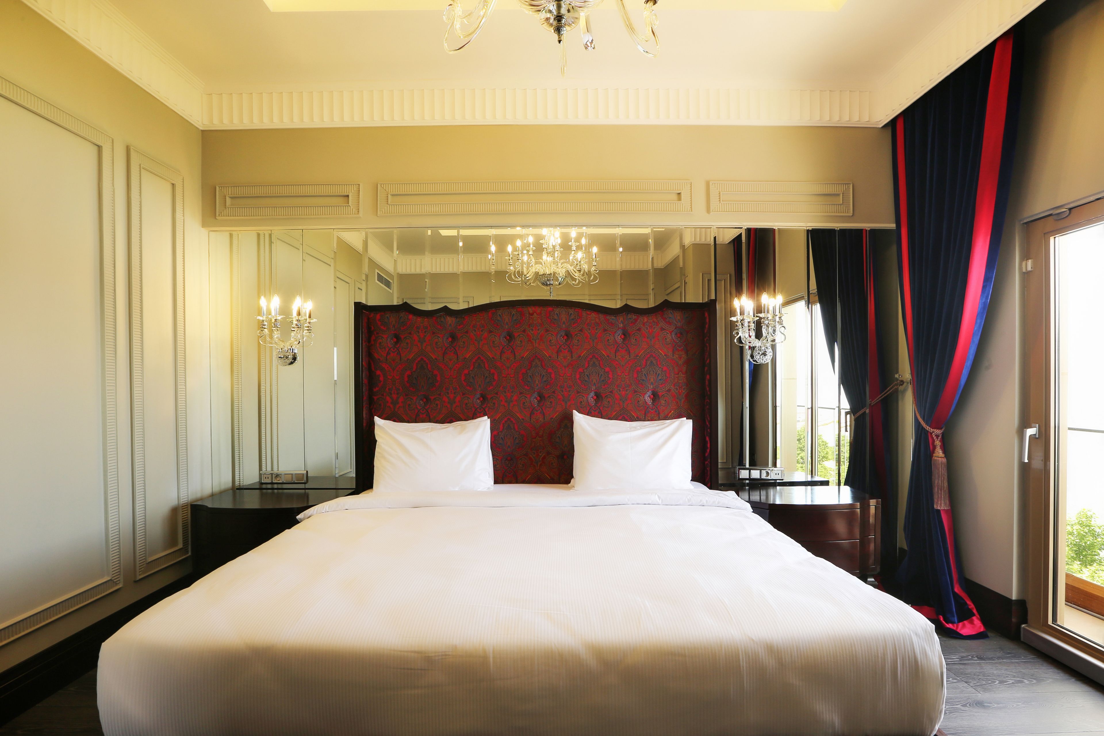 A large bed with white linens, a chandelier, and a red patterned headboard.