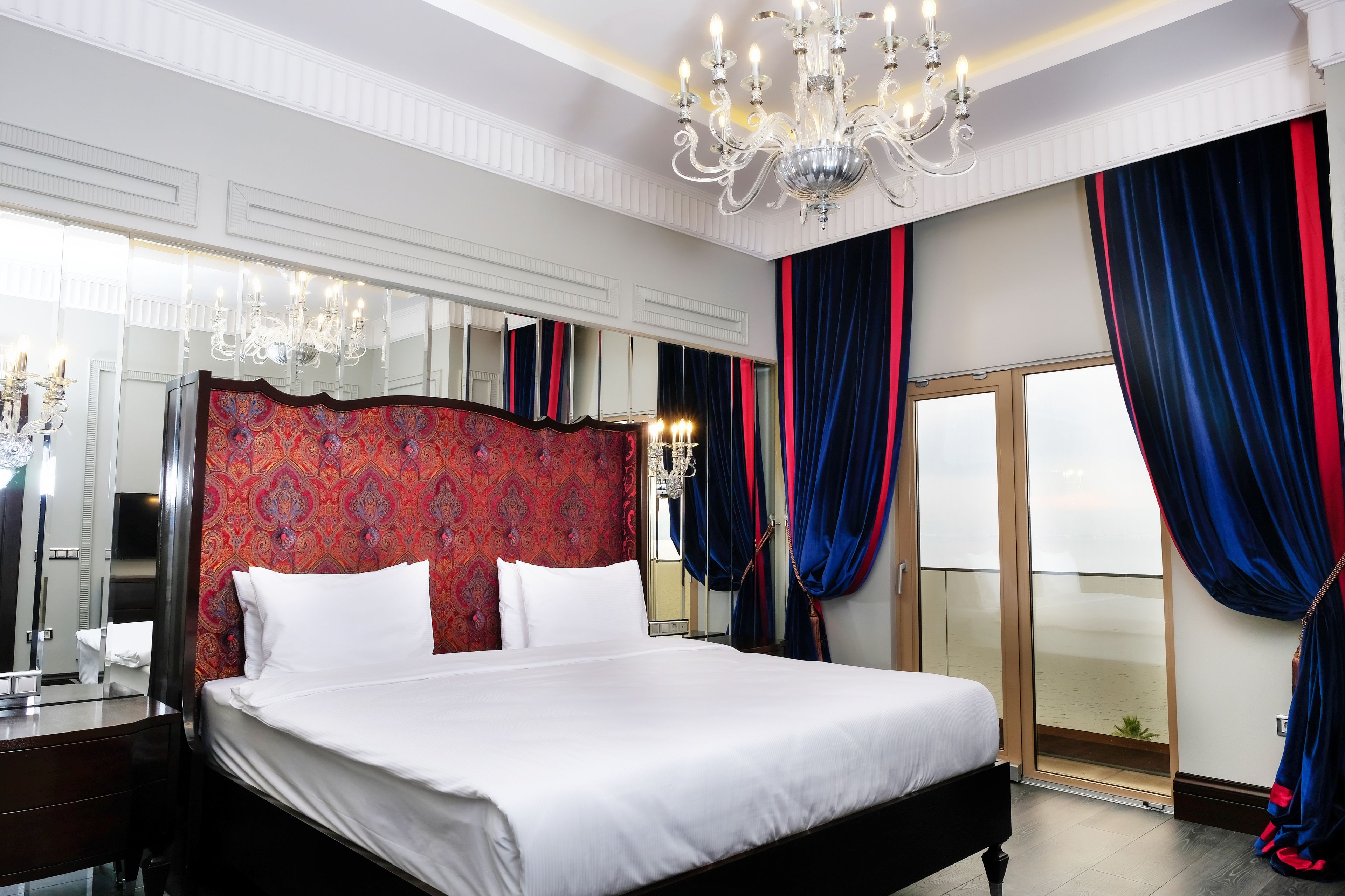 A spacious bedroom with a large bed, a chandelier, a mirror, and a sliding glass door to a balcony.