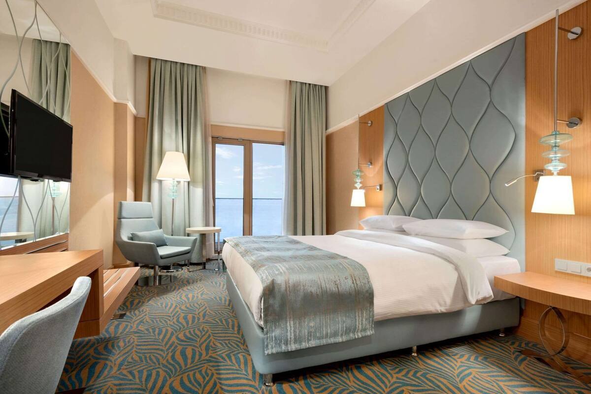 A hotel room with a bed, a desk, a chair, a TV, and a view of the sea.