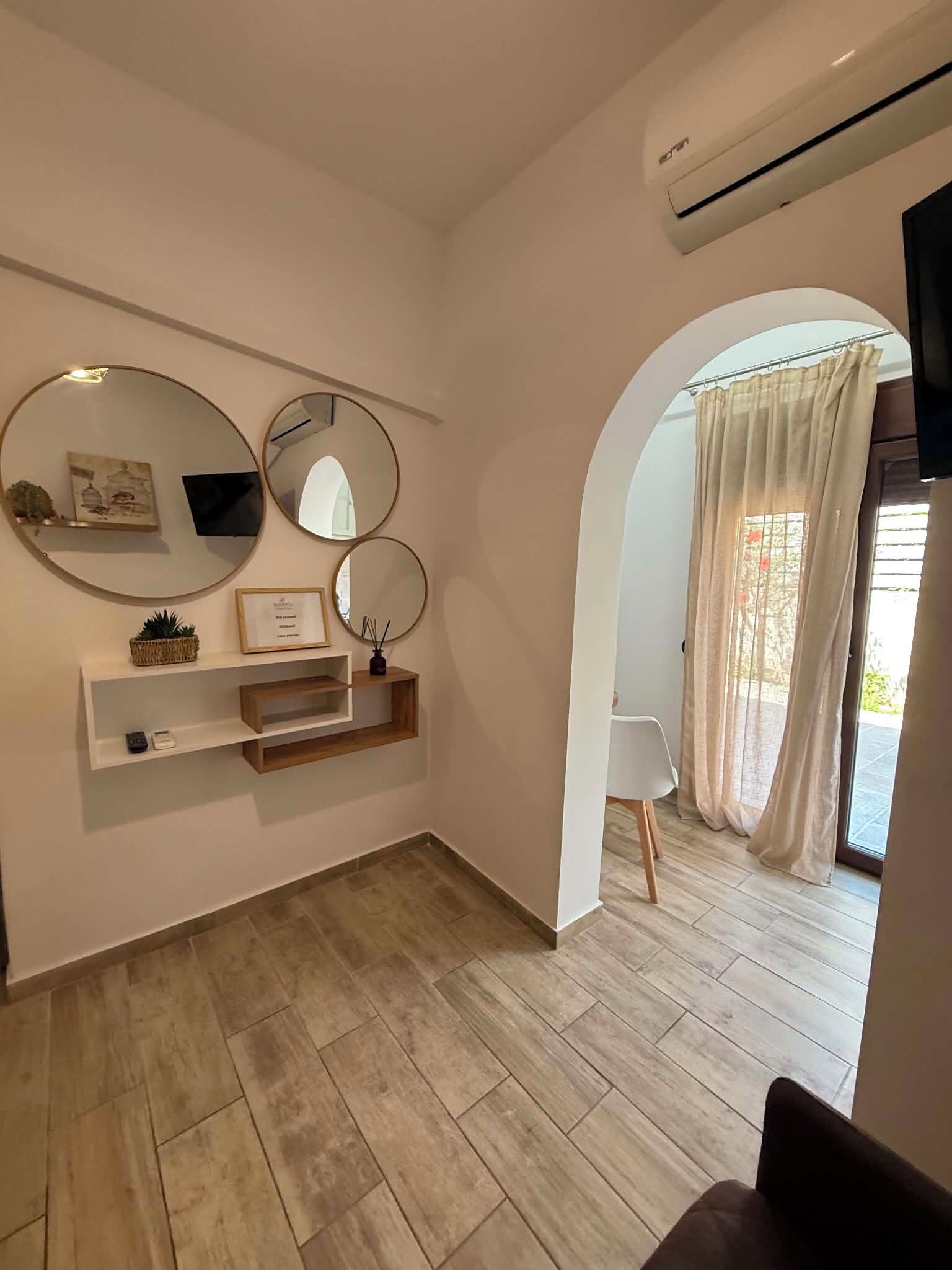 A modern interior with a wooden floor, a white console table with mirrors, a framed picture, and a small plant. An arched doorway leads to a balcony with a view.
