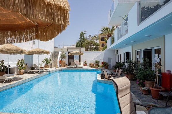 A swimming pool with lounge chairs and umbrellas, surrounded by potted plants and a building with balconies. - Kasteli Studios & Apartments (Mylopotamos)