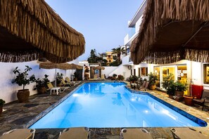 A swimming pool with lounge chairs and potted plants, surrounded by buildings with thatched roofs. - Kasteli Studios & Apartments (Mylopotamos)