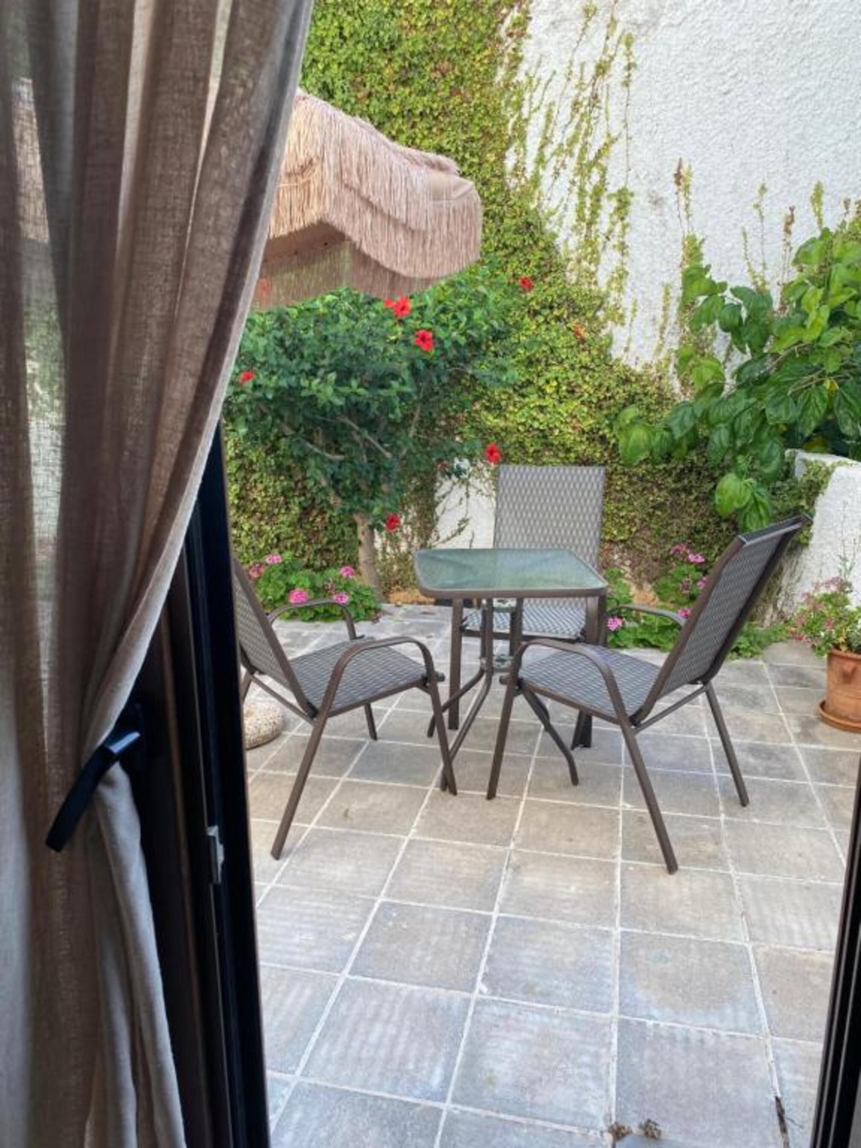 Un patio con mesa y sillas, rodeado de un jardín con diversas plantas y flores.