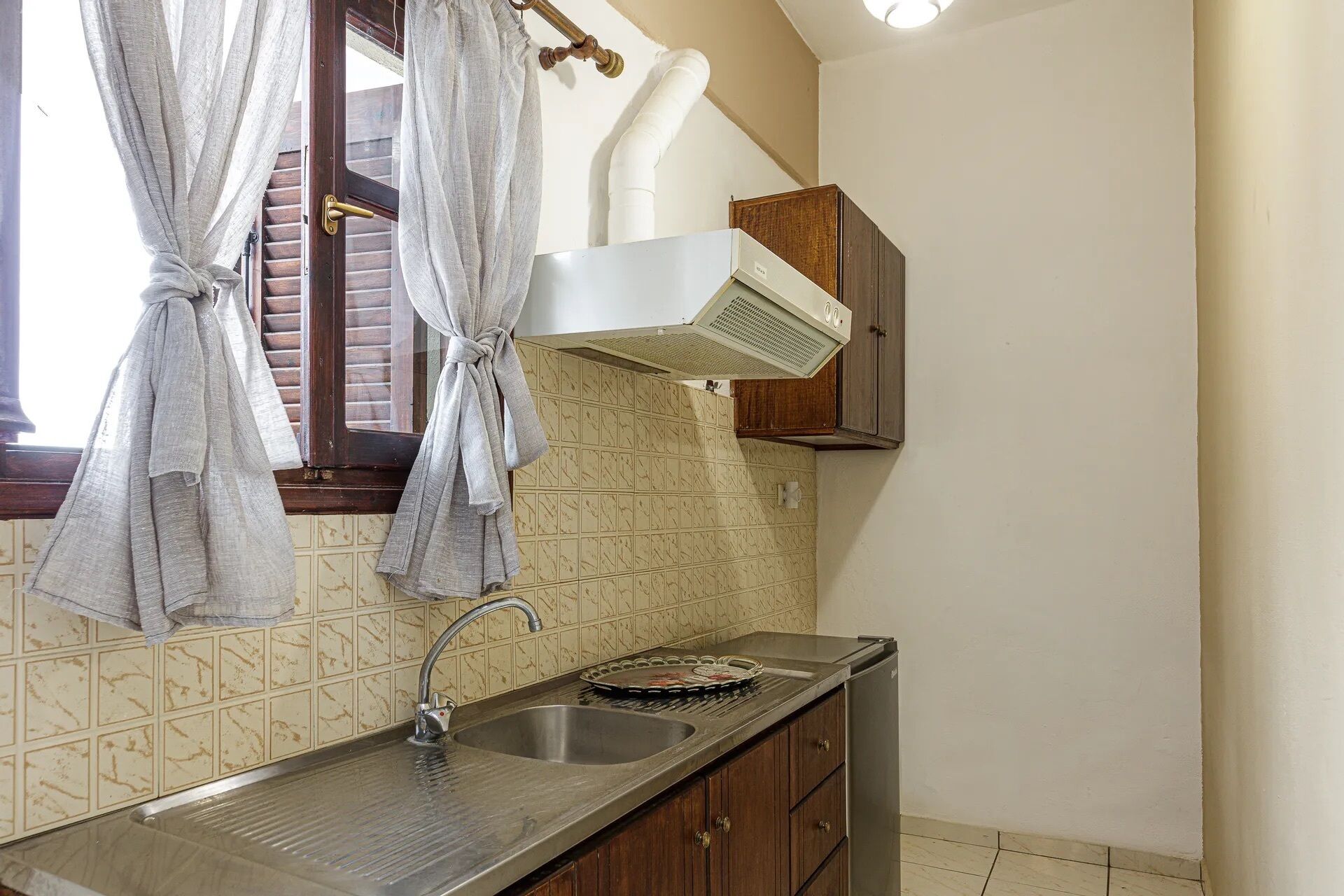 Una cocina con un lavamanos, una ventana con cortinas, una campana extractora y paredes con azulejos.