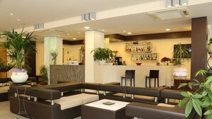 A modern lobby with a sectional sofa, a reception desk, and a bar area with shelves of bottles. - Hotel Ambra (Cesenatico)