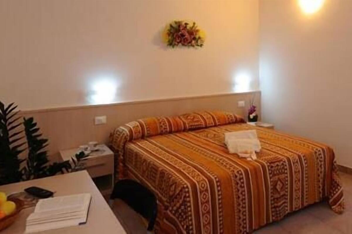 A hotel room with a bed, a nightstand, a desk with a chair, a potted plant, and a wall-mounted light fixture.
