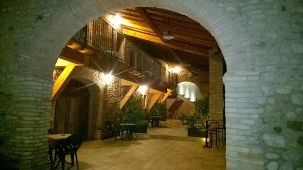 Interior con paredes de piedra, vigas de madera y una escalera que lleva a un nivel superior. Mesas y sillas dispuestas para comer.
