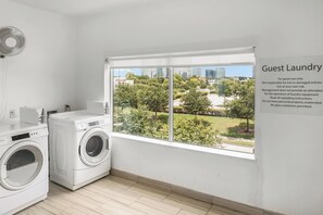 Uma lavanderia para hóspedes com duas máquinas de lavar, um ventilador e uma janela grande com vista para um parque e edifícios.