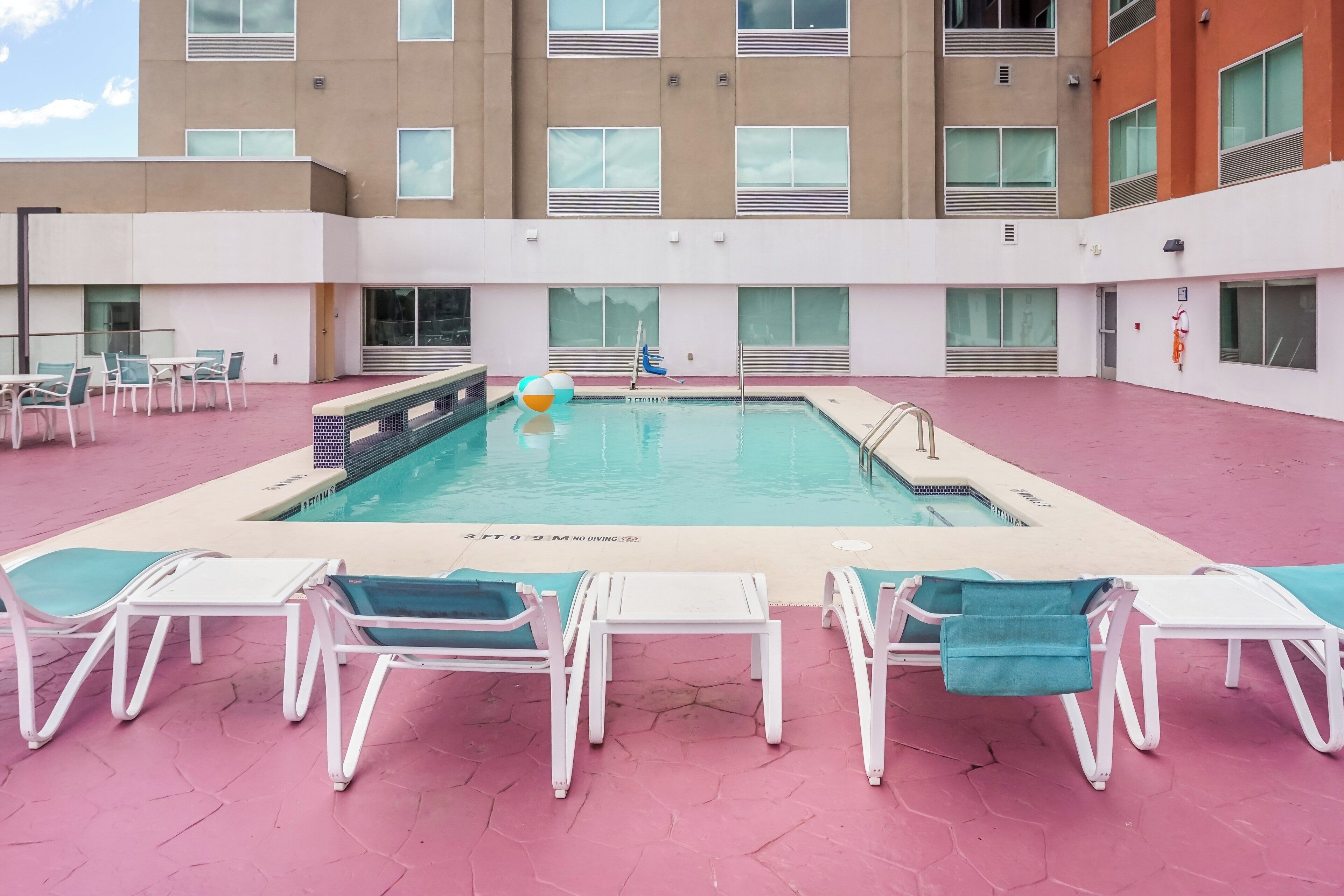 A swimming pool with lounge chairs and a diving board, surrounded by a building with multiple windows.