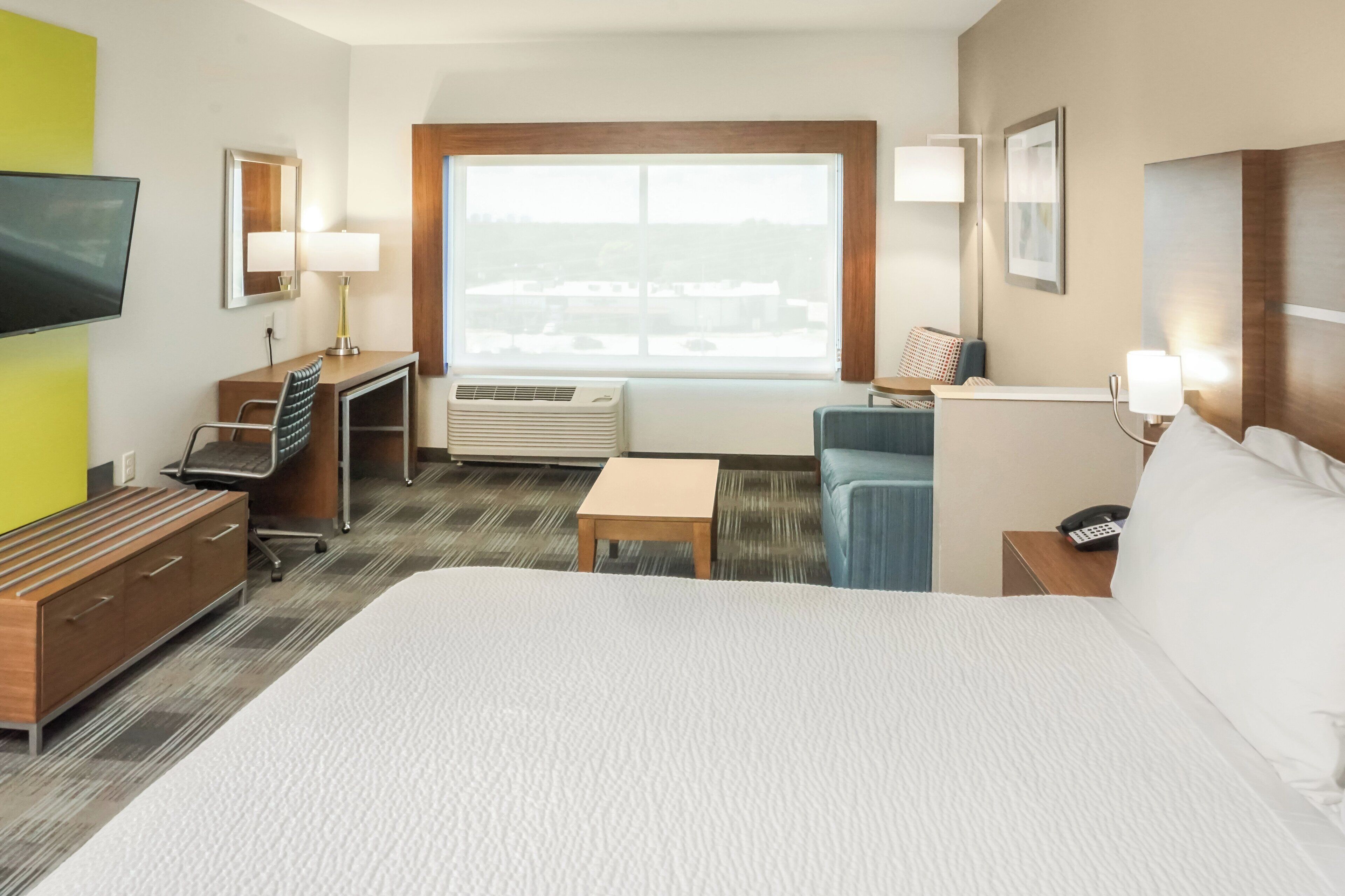 A hotel room with a large bed, a desk with a chair, a small table, a TV, and a window with a view of buildings.