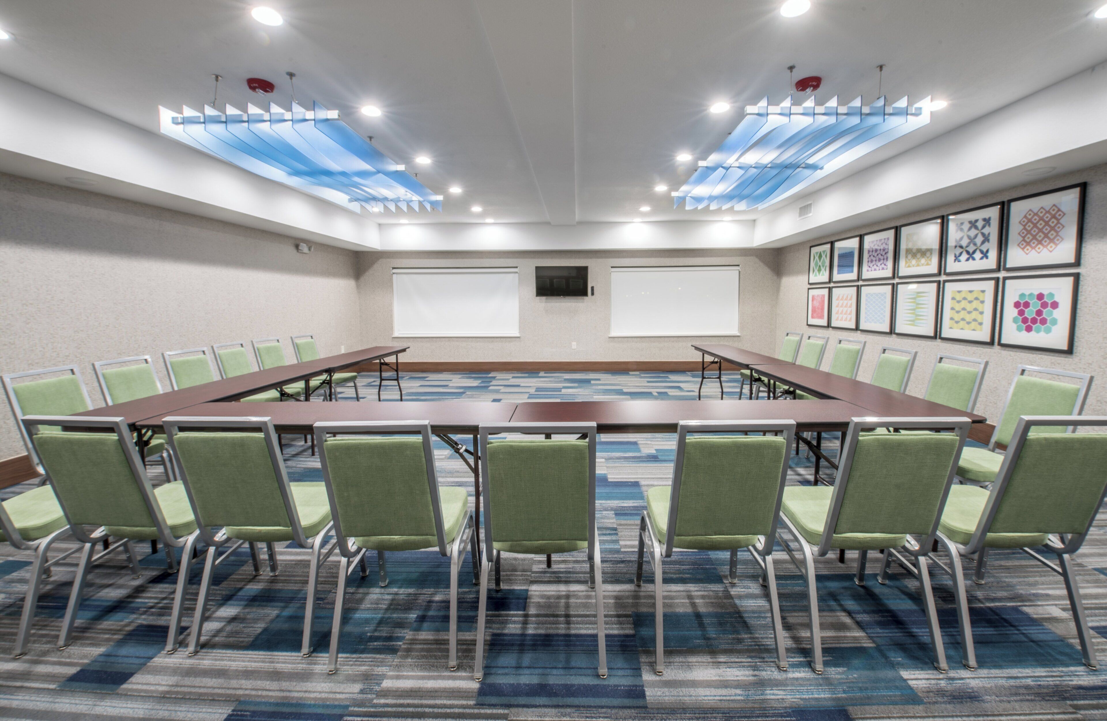 A conference room with a long table, green chairs, white screens, and patterned carpet.