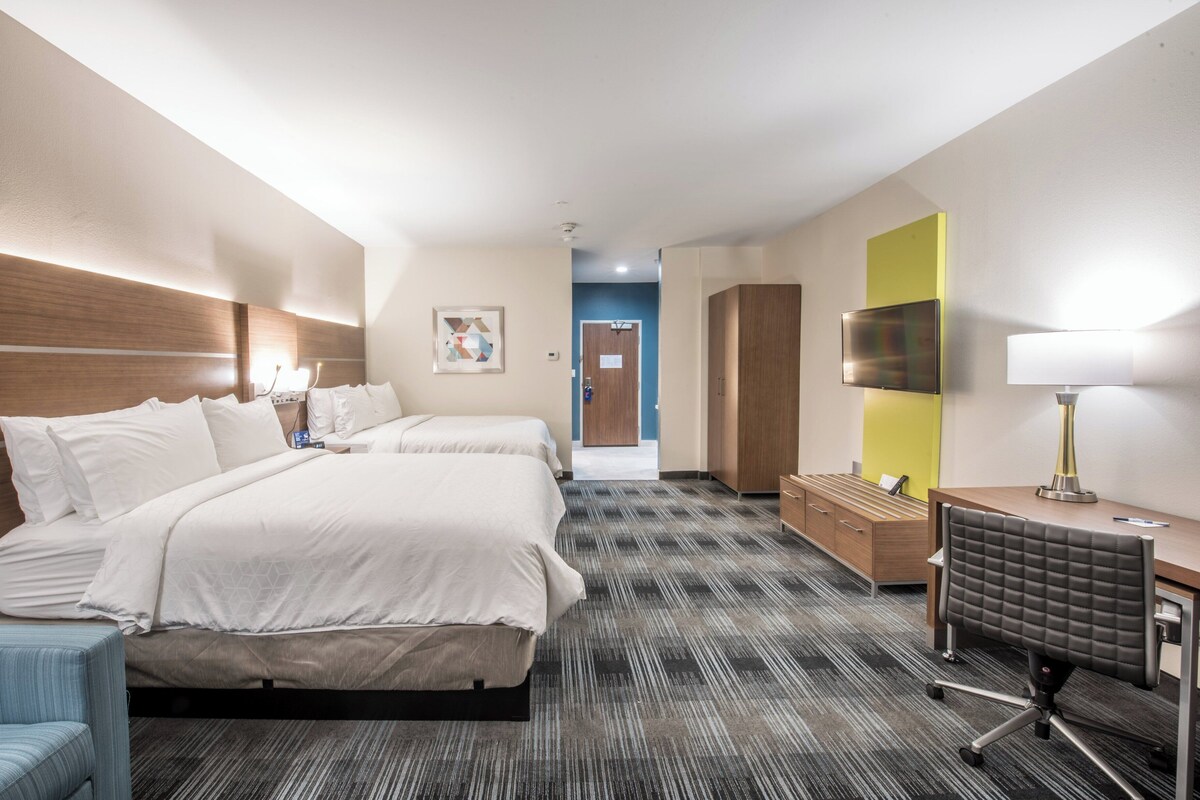 Quarto de hotel com duas camas, uma escrivaninha, uma cadeira, televisão e porta de banheiro ao fundo.