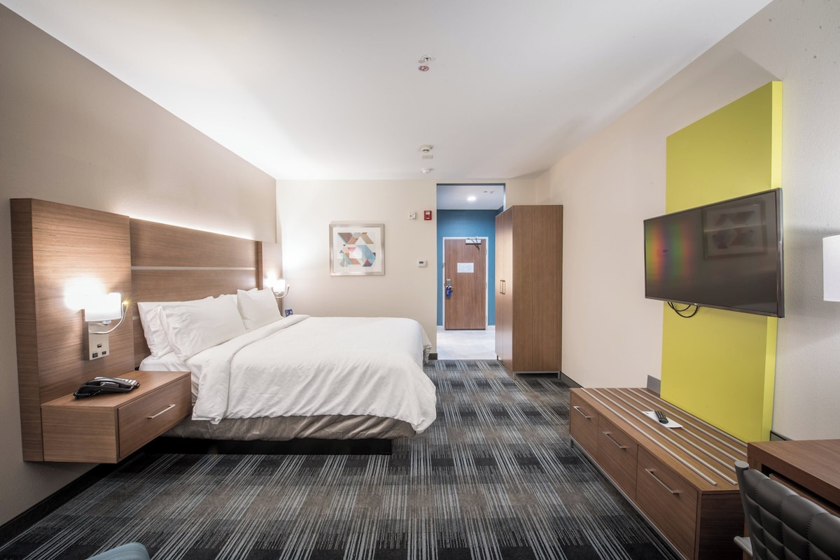 Quarto de hotel com cama, mesa de cabeceira, luminária, televisão e escrivaninha com gavetas.