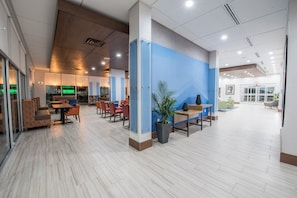 Um hall espaçoso com design moderno, apresentando uma área de estar com mesas e cadeiras, uma grande planta em vaso e uma parede azul de destaque.