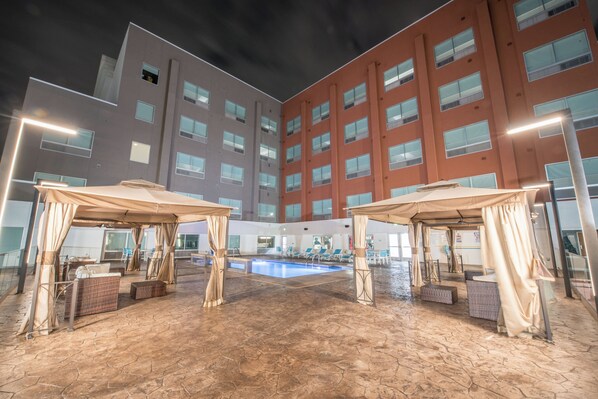 Pátio de hotel com piscina, área de estar externa e um edifício grande ao fundo.