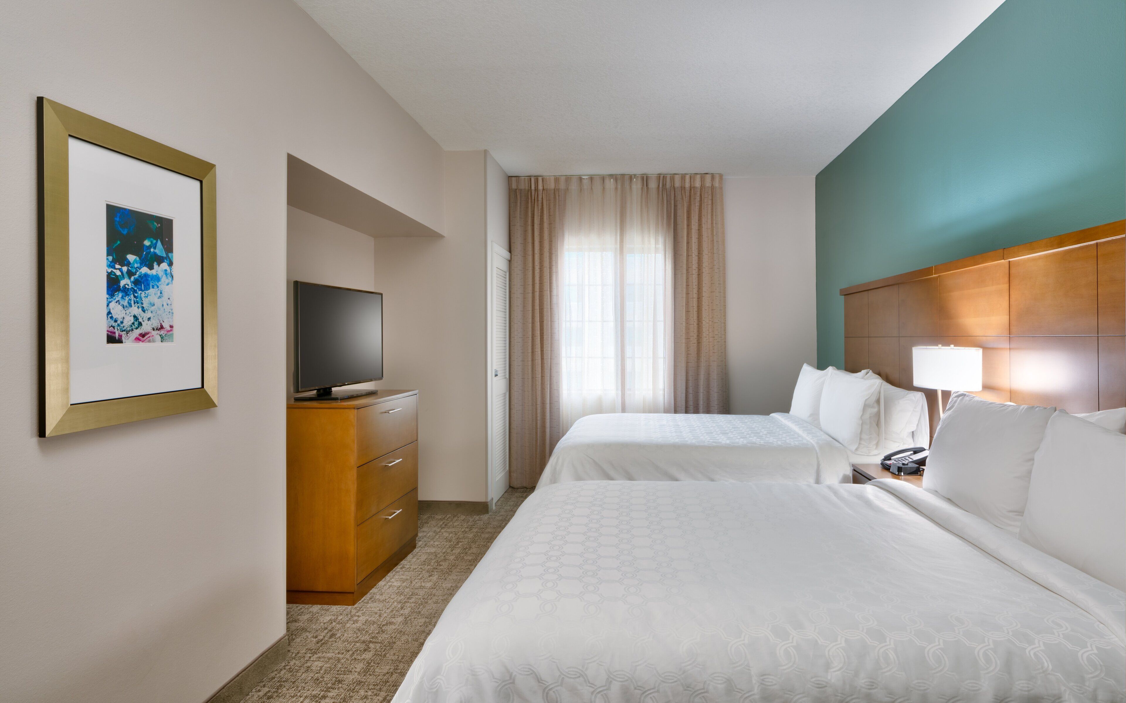 A hotel room with two beds, a television on a wooden dresser, a framed picture on the wall, and a window with curtains.