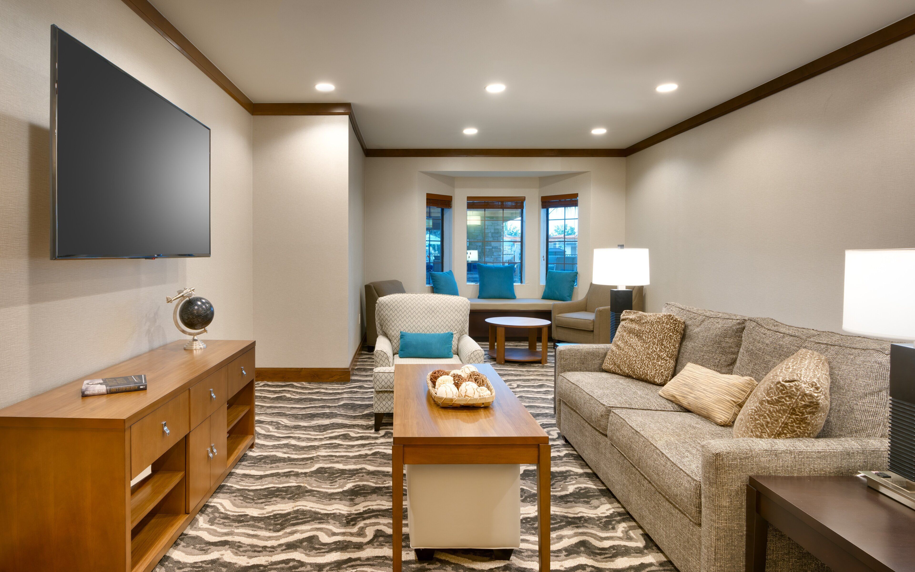 A hotel room with a flat-screen TV, a wooden side table, a patterned rug, a sofa with cushions, and a window with a view.