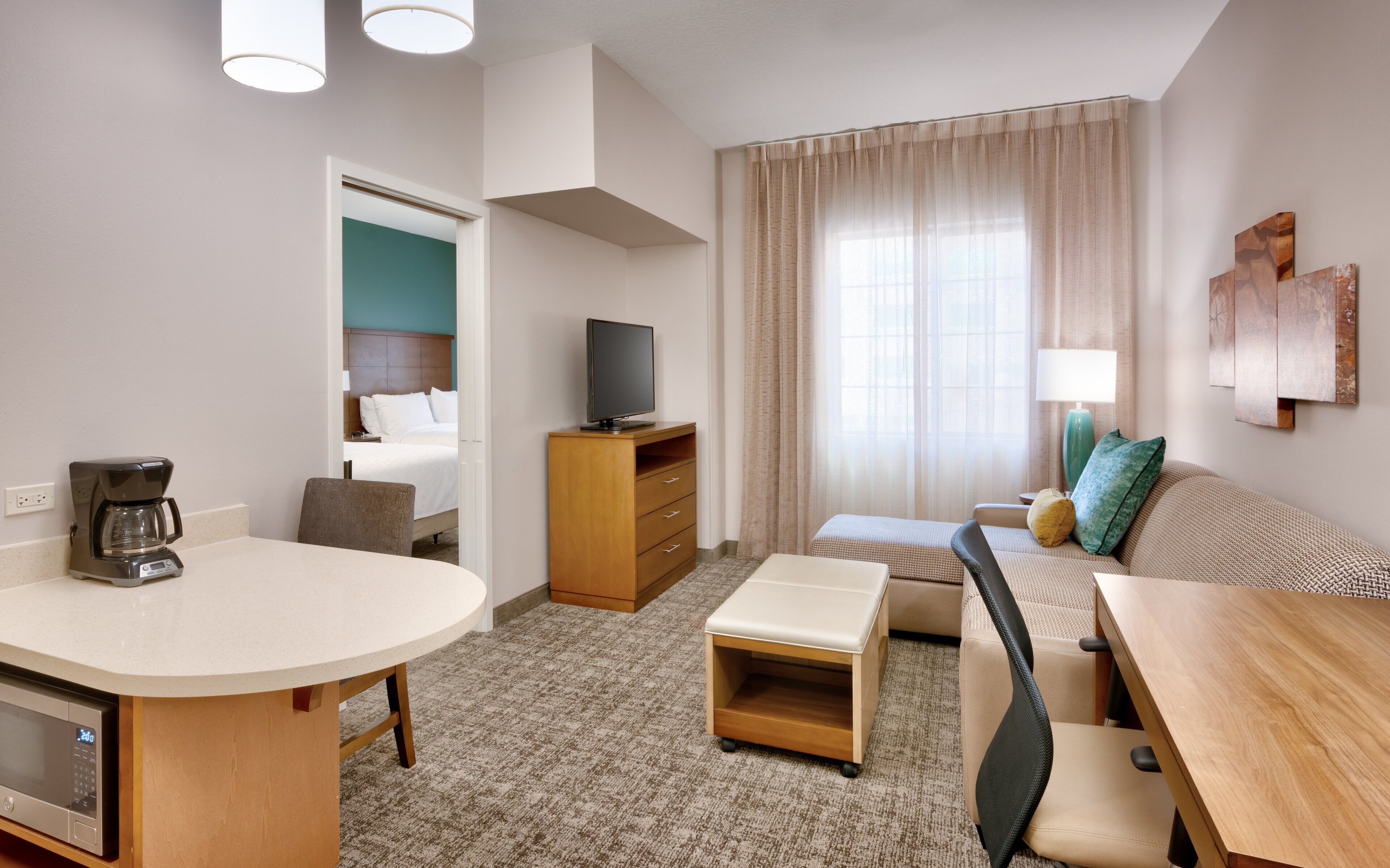 A hotel room with a bed, a television, a desk with a coffee maker, a sofa, and a window with curtains.