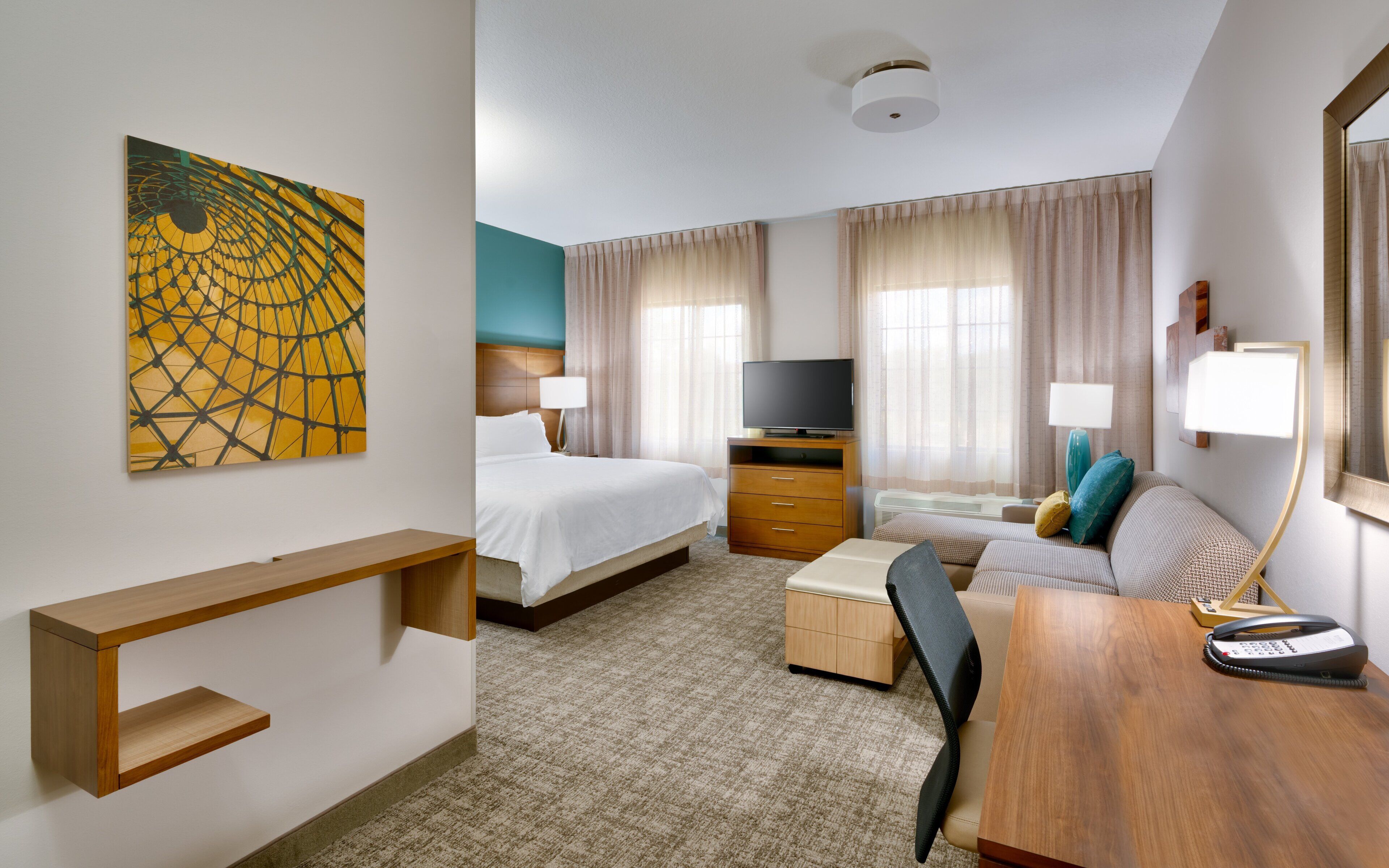 A hotel room with a bed, a desk, a chair, a television, and a painting on the wall.
