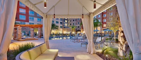 A covered outdoor seating area with a pool and lounge chairs, surrounded by buildings.