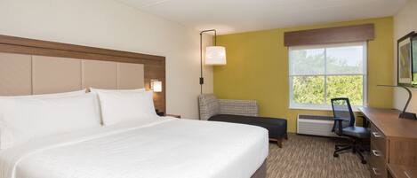 A hotel room with a large bed, a desk, a chair, and a window with a view of greenery.