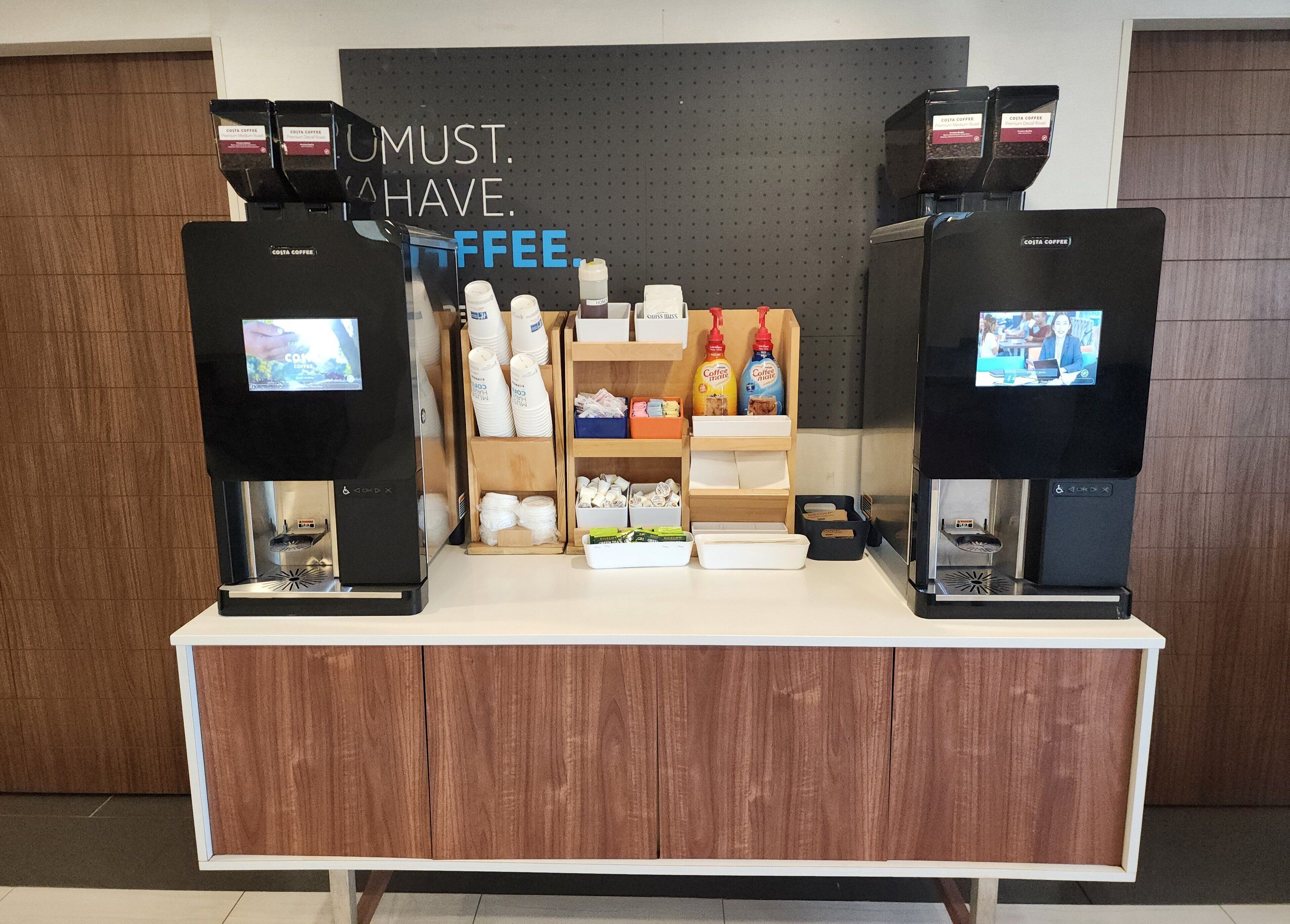 Un espace dédié au café, avec deux machines à café, un panneau indiquant « VOUS DEVEZ ABSOLUMENT PRENDRE UN CAFÉ », et divers articles liés au café disposés sur des étagères.