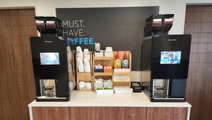 A coffee station with two coffee machines, a sign reading "UMUST. HAVE. COFFEE.", and various coffee-related items on shelves.