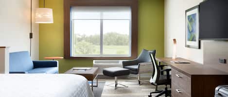 A hotel room with a bed, a desk with a chair, a lamp, and a large window offering a view of trees.