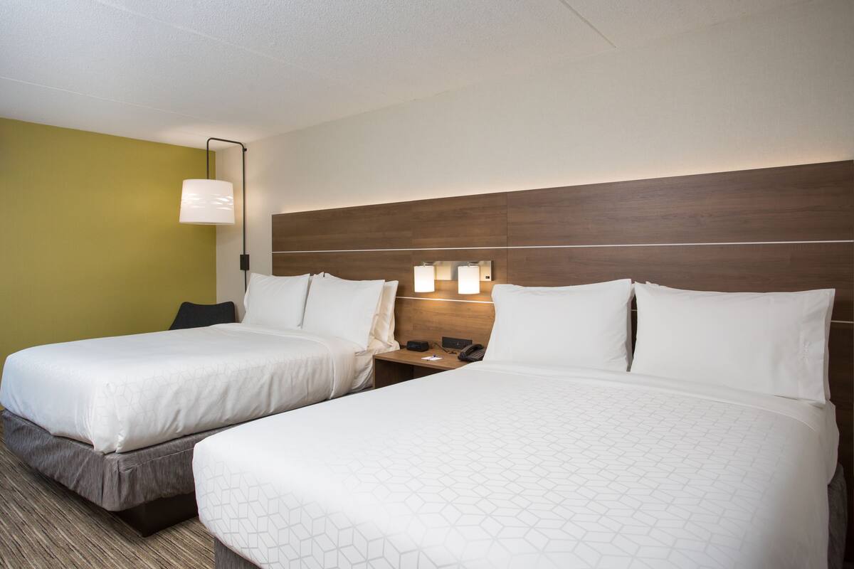A hotel room with two beds, a wooden headboard, a nightstand with a lamp, and a wall-mounted light fixture.