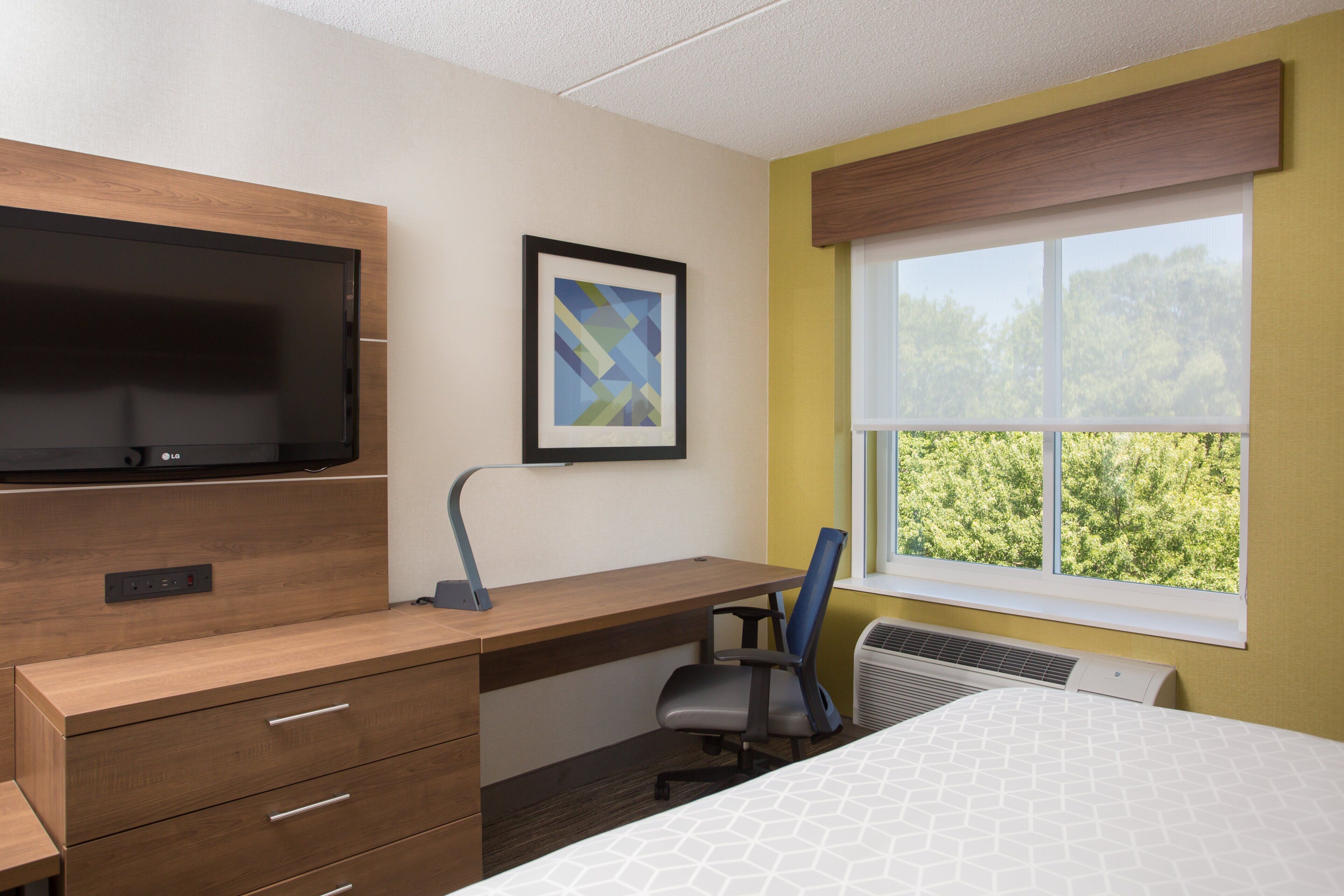 A hotel room with a wooden desk, a flat-screen TV, a framed abstract artwork, a window with a view of greenery, and a bed with a white geometric-patterned bedspread.