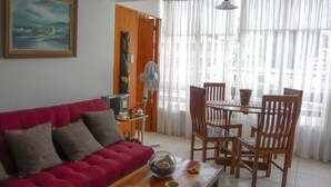 A living room with a red sofa, wooden dining table, and chairs. There is a painting on the wall, a fan, and a window with curtains.