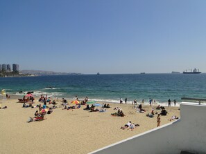 A beach with people sunbathing and swimming, a clear sky, and ships in the distance. - Departamento Reloj De Flores (Vina del Mar)
