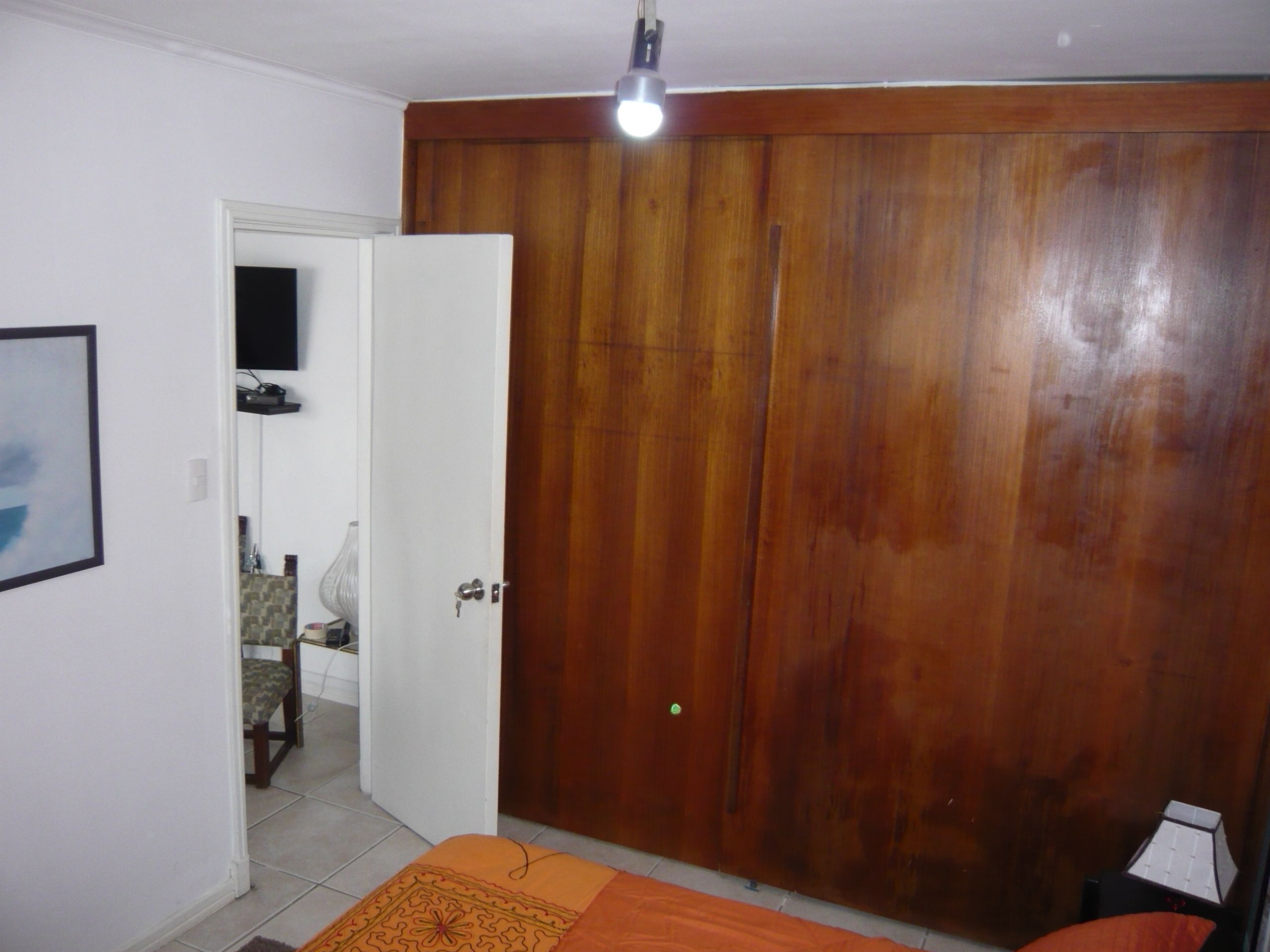 Un dormitorio con un armario de madera, una cama con manta naranja, una puerta blanca y una lámpara en el techo.
